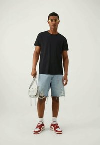 Fekete rövid ujjú póló, világoskék, kopott farmer rövidnadrág, piros és fehér sportcipő, egy kis fehér táskát tart. Átlátszó keretes napszemüveg.