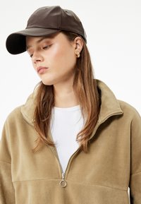 Brun læderhat bæres med en beige fleece jakke med kvart lynlås over en hvid skjorte. Jakken har en sølv lynlås og en blød tekstur.