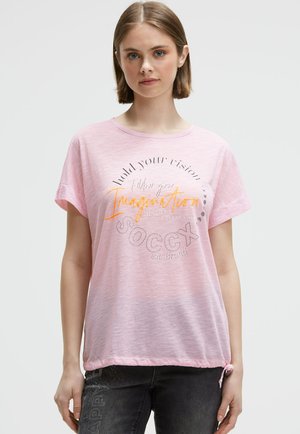 Eine junge Frau mit kurzen braunen Haaren trägt ein hellrosa T-Shirt mit hochgekrempelten Ärmeln und buntem inspirierendem Text, kombiniert mit dunklen Jeans.