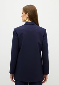 Námořnické sako s přiléhavým střihem, klopovými límečky a dlouhými rukávy. Model je zobrazen zezadu, má volné vlasy a nosí velké žluté kruhové náušnice.