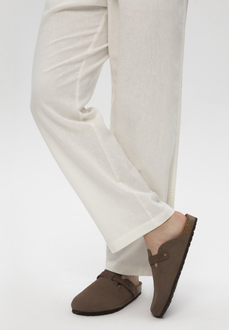 Personne portant un pantalon beige à jambes larges et des sabots marron à enfiler avec détail de boucle, debout sur un pied en croisant l'autre jambe.