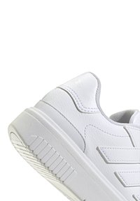 Sneaker blanche en cuir texturé avec une semelle en caoutchouc plate, présentant des coutures discrètes et un logo en relief à l'arrière.