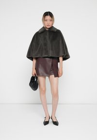 Dunkelgrauer Kunstpelz-Cape mit Kragen, getragen über einem bordeauxfarbenen Leder-Minisrock, kombiniert mit schwarzen spitzen Ballerinas und einer kleinen schwarzen Tasche.