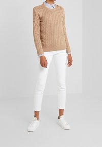 Suéter beige de punto trenzado con escote en V, que cuenta con tres botones dorados en un hombro. Combinado con pantalones ajustados blancos y zapatillas blancas.