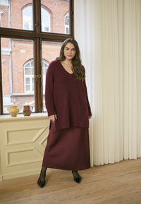 Maglione a maglia bordeaux con scollo a V profondo, abbinato a una lunga gonna coordinata. Tessuto strutturato e ballerine nere a punta completano l'outfit.