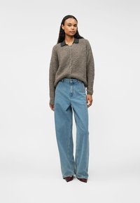 Καφέ υφασμάτινο κασκόλ με κουμπιά, μαύρο γιακά, σε συνδυασμό με ανοιχτό μπλε ψηλόμεσα φαρδιά τζιν και σκούρα παπούτσια, στέκεται σε ένα απλό φόντο.