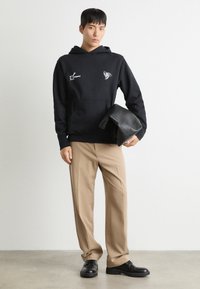 Sweat à capuche noir avec des détails graphiques blancs, pantalon beige et chaussures noires. Le modèle tient un sac en cuir noir. Fond en béton.