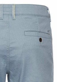 Lichtblauwe katoenen broek met een gladde textuur, voorzien van een achterzak met een knop. Gele stiksels accentueren de tailleband.
