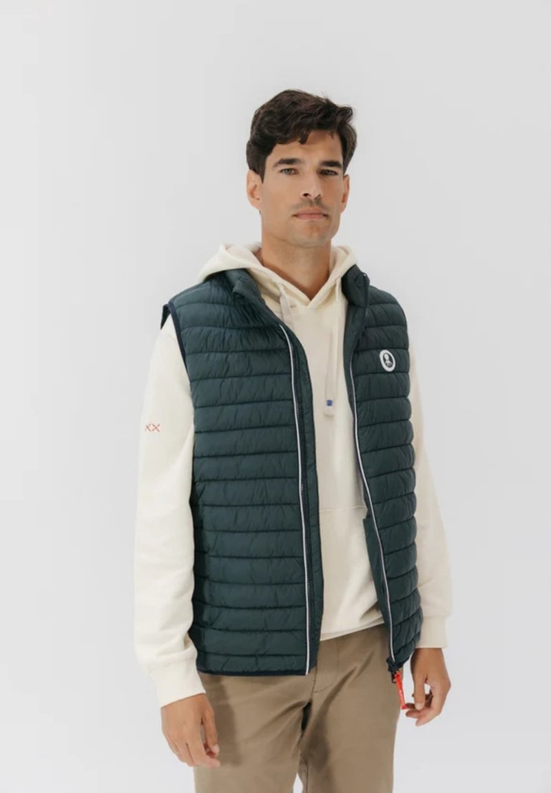 Chaleco acolchado verde con costuras horizontales, cierre con cremallera y un logo. Llevado sobre una sudadera ligera con capucha, combinado con pantalones beige. Diseño sencillo.