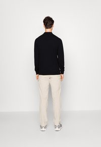 Haut noir à manches longues, coupe ajustée. Pantalon crème avec poches latérales. Le modèle porte des baskets grises. Fond uni.