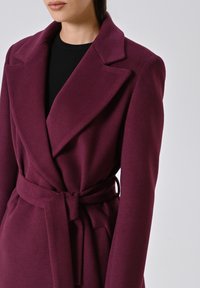 Burgunderfarbener Woll-Mix-Mantel mit Reverskragen, Bindegürtel und strukturiertem Stoff. Besitzt lange Ärmel und eine taillierte Silhouette.