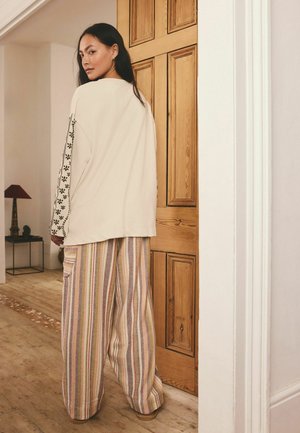 Žena stojí v miestnosti pri drevených dverách, má na sebe krémový top s čiernymi kvetinovými rukávmi a široké pásikavé nohavice.