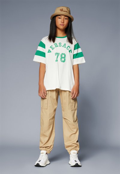 Valge T-särk roheliste triipudega ja "VERSACE 78" disain, koos beeži cargo-pükstega. Jalatsiteks on valged tossud mustade aktsentidega.