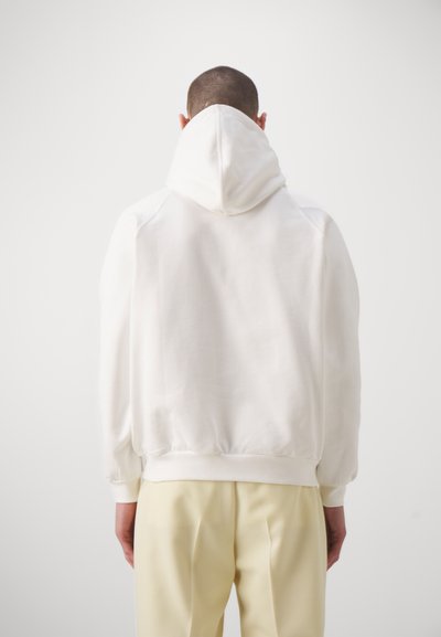 Felpa bianca con cappuccio, polsini e fondo a costine. Il tessuto sembra morbido. Indossata con pantaloni giallo chiaro, che mostrano un fit rilassato.