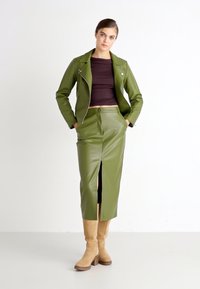 Veste en simili cuir vert olive avec des accessoires en argent, superposée à un haut marron, assortie à une jupe et des bottes beiges jusqu'au genou.