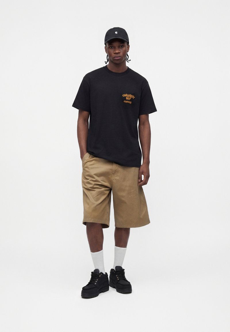 Mann steht, trägt eine schwarze Mütze, ein schwarzes T-Shirt mit Grafik, beige knielange Shorts, weiße Crew-Socken und schwarze Sneaker, Hände in den Taschen.
