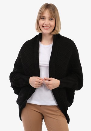 Donna che sorride, indossando un poncho nero a trama sopra una camicia bianca e pantaloni beige, in piedi contro uno sfondo chiaro e semplice.