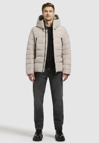 Lys grå polstret jakke med hætte, der har lynlås lommer, en front lynlås, og en glat overflade; sat sammen med mørke jeans og sorte sko.