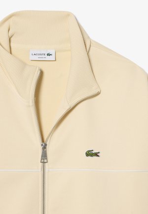 Sweat-shirt Lacoste beige à coupe ample avec fermeture éclair, col côtelé, fermeture éclair argentée et logo crocodile vert sur la poitrine.
