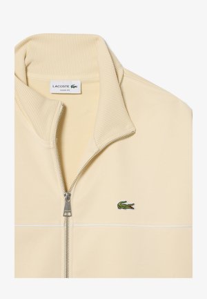 Sweat-shirt Lacoste beige à coupe ample avec fermeture éclair, col côtelé, fermeture éclair argentée et logo crocodile vert sur la poitrine.