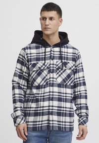 Camicia flanella a quadri in blu navy e bianco, con cappuccio nero, due tasche sul petto e chiusura con bottoni, maniche lunghe e vestibilità rilassata.