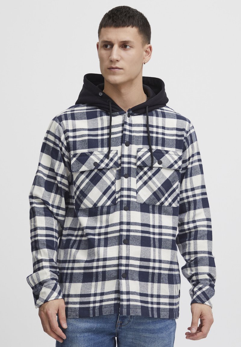Camicia flanella a quadri in blu navy e bianco, con cappuccio nero, due tasche sul petto e chiusura con bottoni, maniche lunghe e vestibilità rilassata.