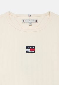 Kreminės spalvos trumpomis rankovėmis megzta marškinėlių su mažu daugiaspalviu „TOMMY HILFIGER“ logotipo lopu ant krūtinės. Minkšta medžiaga ir apvali iškirptė.