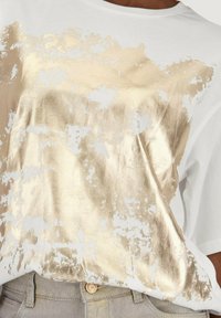 Witte T-shirt met een vergrijsde gouden metallic print, met een ronde hals en korte mouwen, gecombineerd met lichtgrijze broek.