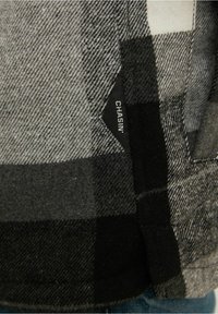 Szara i czarna flanelowa tkanina w kratę o teksturowanej powierzchni; posiada czarną metkę z napisem "CHASIN" na bocznym szwie.