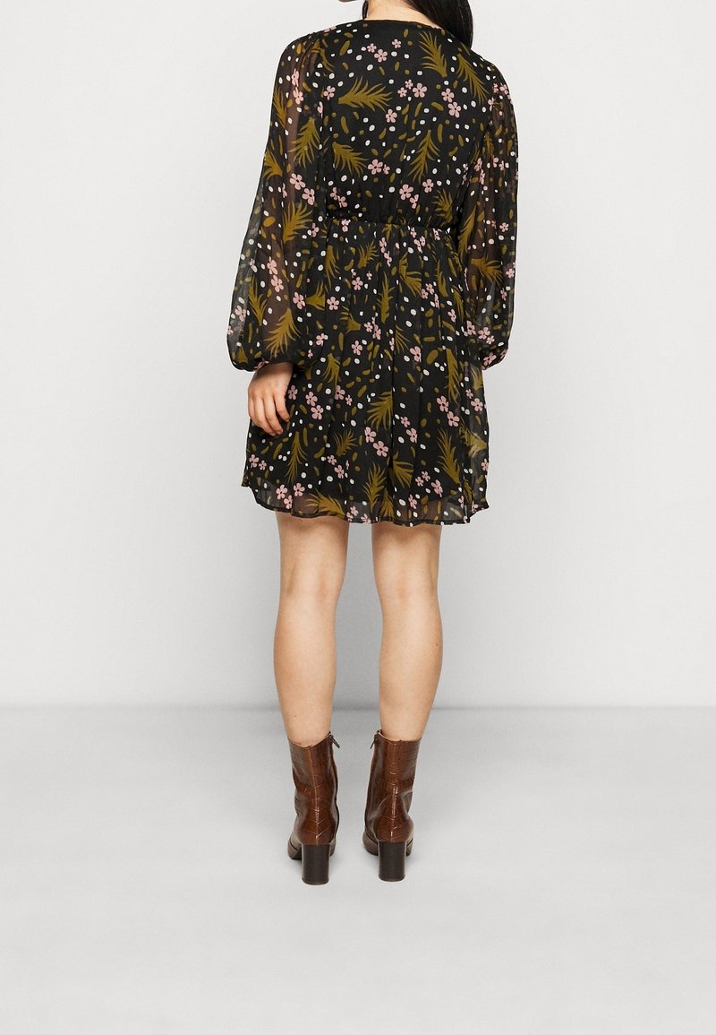 Černé květinové šaty s dlouhými, průhlednými rukávy, zdobené růžovými květy a zelenými listy, stažené v pase. Hnědé boty na podpatku dokončují vzhled.