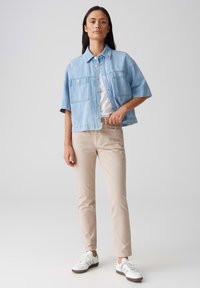 Lichtblauwe denim korte mouwen jacket met twee borstzakken, gedragen over een wit T-shirt, gecombineerd met beige slim-fit broek en witte sneakers.