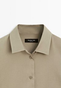 Camicia beige con colletto a punta, due bottoni visibili e un'etichetta nera con la scritta "Massimo Dutti, Made in China."