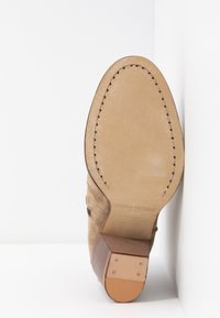 Brązowa zamszowa podeszwa z gładkim skórzanym wykończeniem, z przyszytym brzegiem i drewnianym obcasem, ustawiona pod kątem na białym tle.