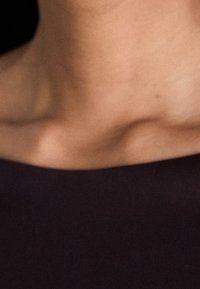 Tmavá tkanina hladce splývá přes ramena, vystavuje čistý výstřih a jemné tóny pleti s měkkou strukturou a teplým osvětlením.