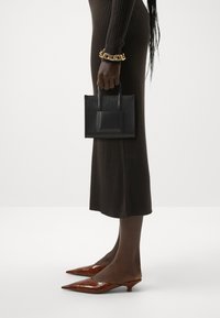 Borsa nera texture con manici corti, tenuta in una mano. Il modello indossa un vestito marrone a coste e scarpe a punta lucide marrone.