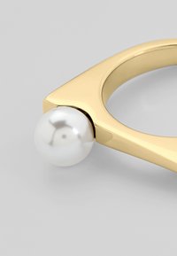 Anello d'oro con design triangolare e finitura lucida, caratterizzato da una perla bianca lucida all'estremità. Texture liscia e dettagli eleganti.
