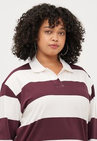 Une jeune femme aux cheveux bouclés et portant des boucles d'oreilles créoles est habillée d'un polo rayé bordeaux et blanc, regardant la caméra avec confiance.