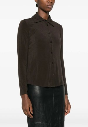 Donna che indossa una camicia marrone scuro a maniche lunghe con colletto largo e una gonna in pelle nera.