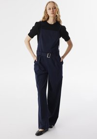 Salopette blu navy con vita regolabile, gambe ampie e tasche applicate, abbinate a una maglietta nera a maniche corte. Texture liscia e design strutturato.