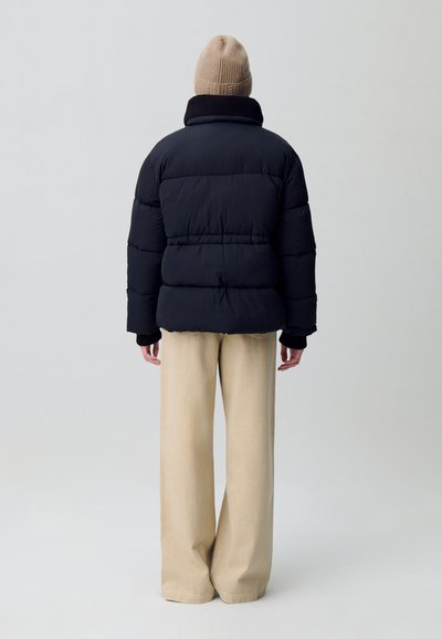 Giacca a vento nera con colletto alto e polsini a coste, abbinata a pantaloni beige a gamba larga. L'outfit è completato da un berretto lavorato a maglia color marrone chiaro.