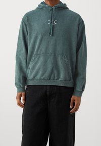 Sudadera color verde azulado hecha de una tela suave, con capucha ajustable, bolsillo tipo canguro y puños acanalados, que presenta un pequeño logo en el pecho.