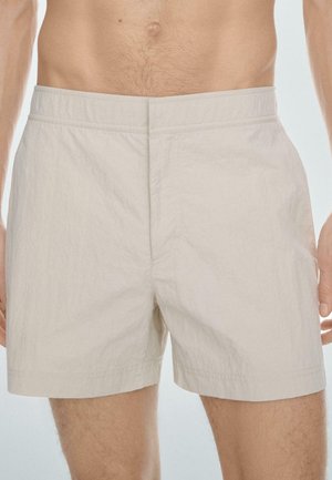 Béžové pánské lehké šortky s elastickým pasem nošené stojícím mužem s viditelným nahým trupem.