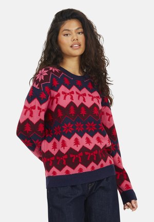Jonge vrouw met lang krullend haar, gekleed in een feestelijke rode, roze en marineblauwe trui met patronen van een kerstboom, strik en sneeuwvlokken.