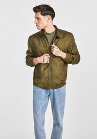 Oliwkowa bomberka z teksturowanym wykończeniem, z zamkiem błyskawicznym i ściągaczami, w połączeniu z jasnoniebieskimi dżinsami.