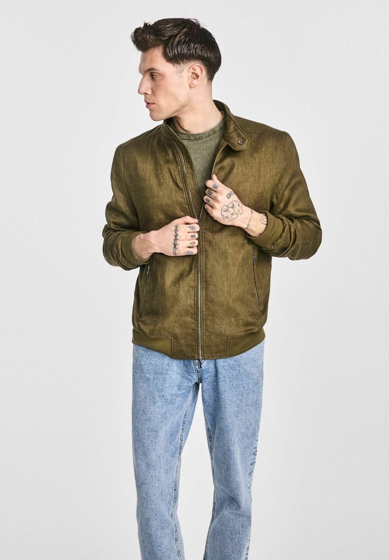 Oliwkowa bomberka z teksturowanym wykończeniem, z zamkiem błyskawicznym i ściągaczami, w połączeniu z jasnoniebieskimi dżinsami.