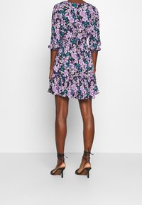 Blumenmusterkleid in Lila, Schwarz und Weiß mit Dreiviertelärmeln und gerafftem Saum. Tragen mit schwarzen Riemchen-Heels. Rückenansicht gezeigt.