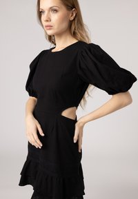 Black dress with puff sleeves, cut-out side detail, and layered ruffled hem. Made of textured fabric with lace accents at the waist.