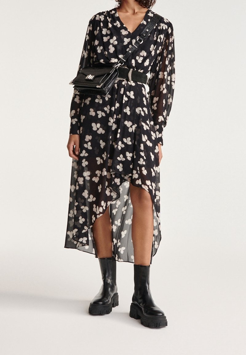 Femme portant une robe asymétrique noire à motif floral, ceinture noire, sac bandoulière et bottines noires épaisses, debout devant un fond uni.