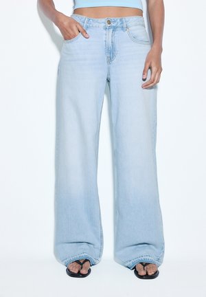 Personne portant un jean large bleu clair, une main dans la poche, associée à des sandales noires à lanières sur fond blanc.