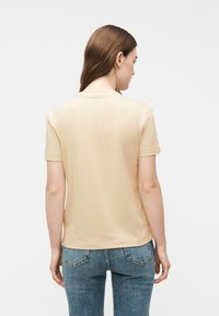 Γυναίκα με μακριά καστανά μαλλιά, που φοράει μπεζ κοντομάνικη μπλούζα και μπλε τζιν, εμφανιζόμενη από πίσω με φόντο ένα απλό φόντο.